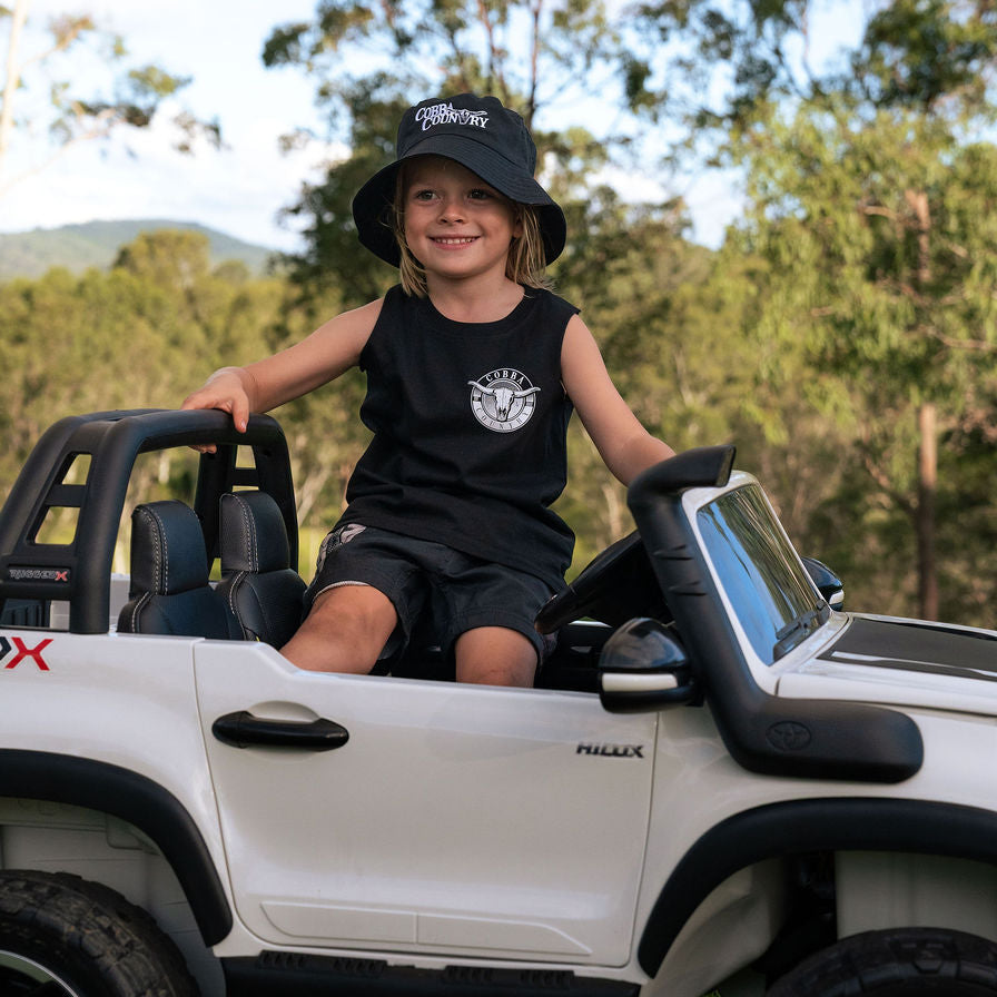 Child sitting in a toy car outdoors with trees in the background wearing Cobba country Tank and Bucket Hat