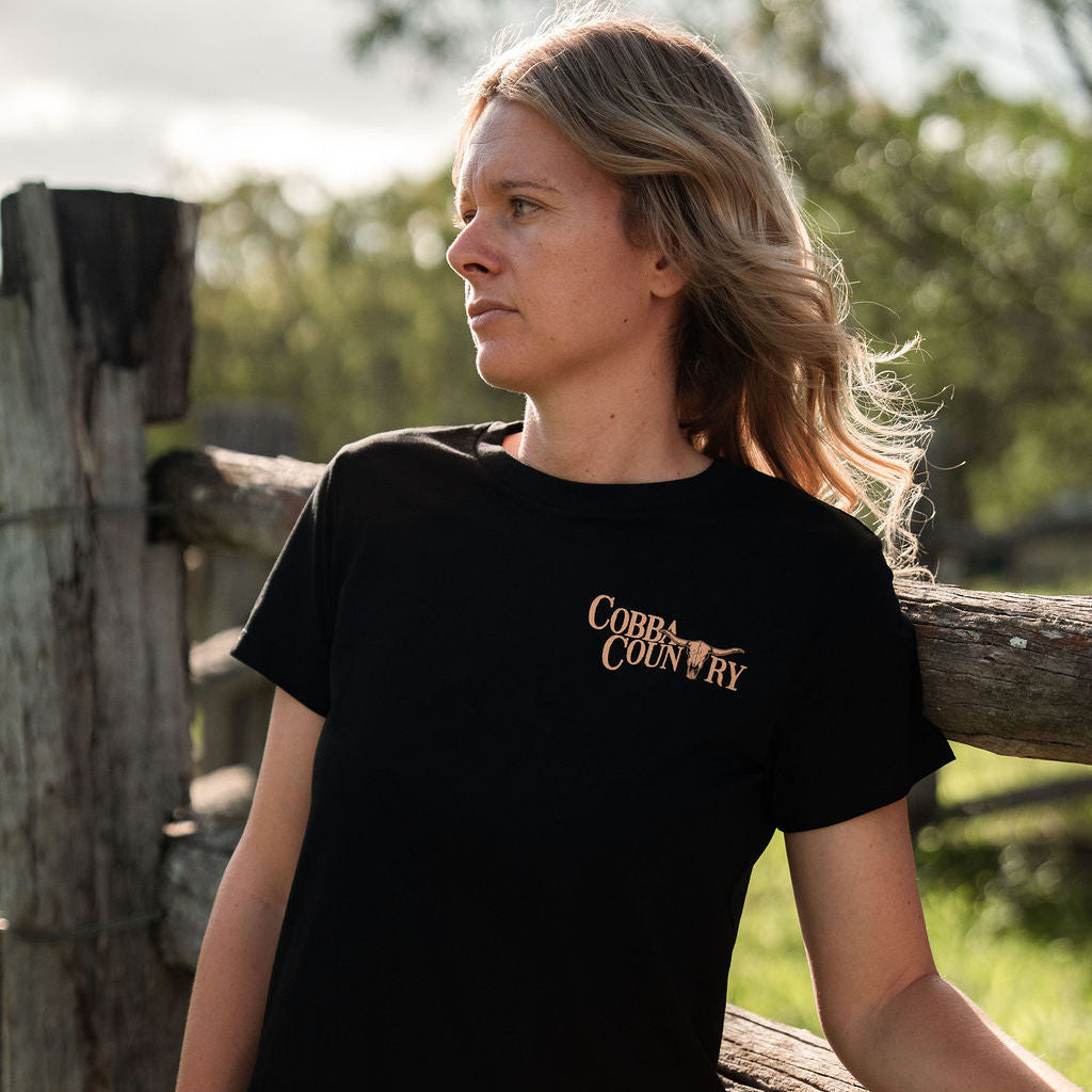 Woman wearing a black t-shirt with 'Coba Country' logo, standing by a wooden fence outdoors.