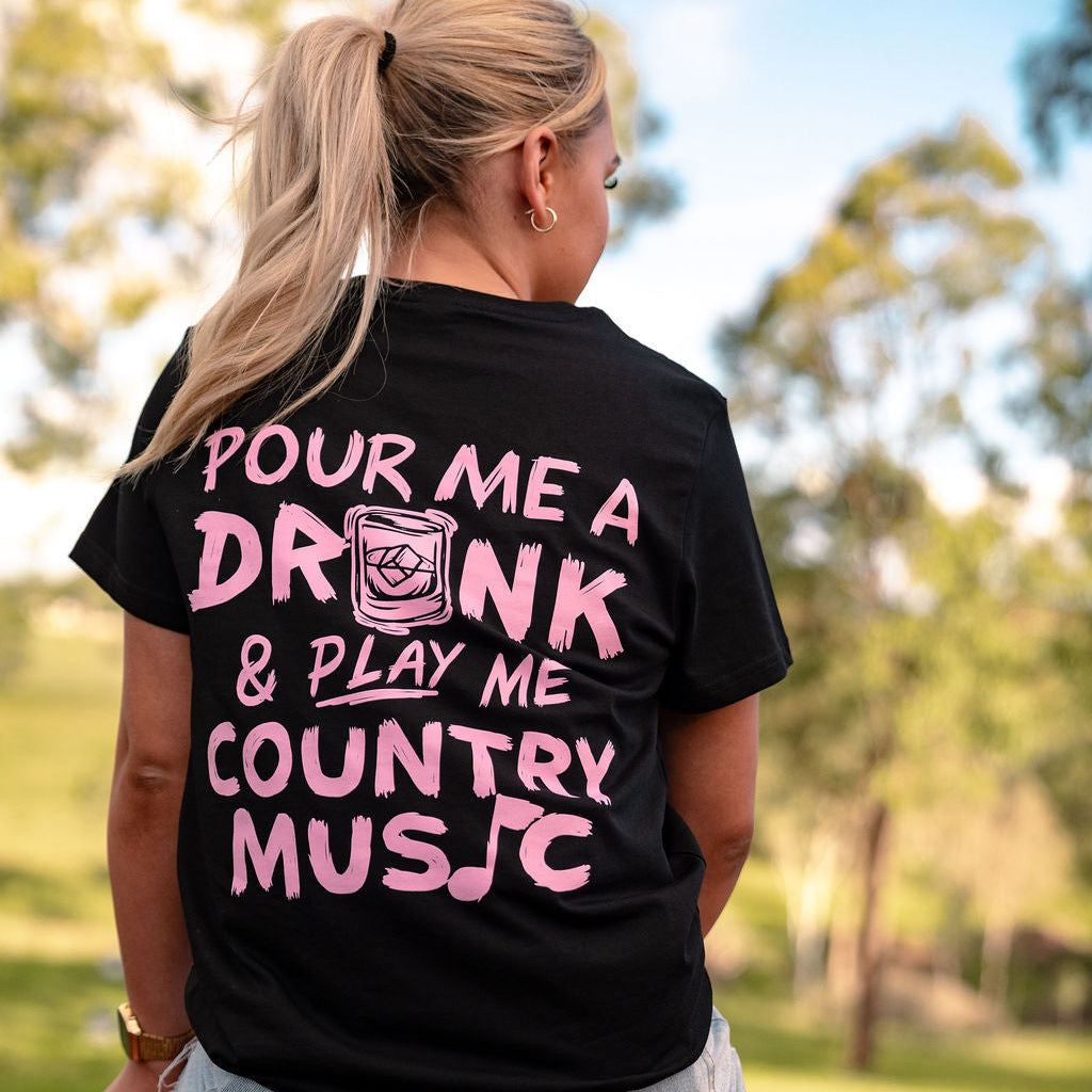 woman sitting on back of ute wearing pour me a drink and play me country music t-shirt